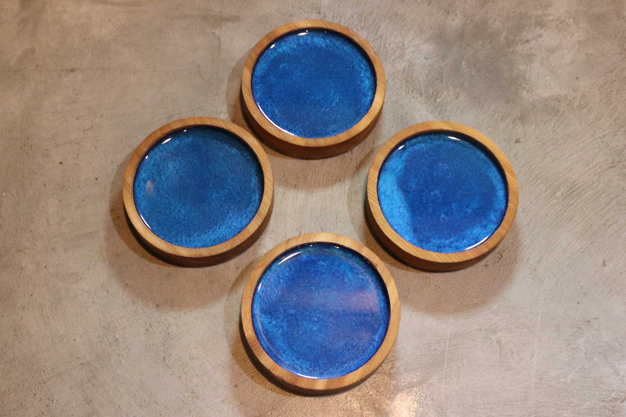 Four Customized Round Cedar Coasters by Willy's Woods, with blue ceramic centers, are elegantly arranged on a concrete surface.