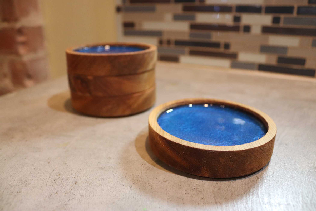 Two Willy's Woods' Customized Round Cedar Coasters, featuring blue ceramic centers, rest on a countertop. One is stacked among others, while the other lies separately. A tiled wall appears behind them.