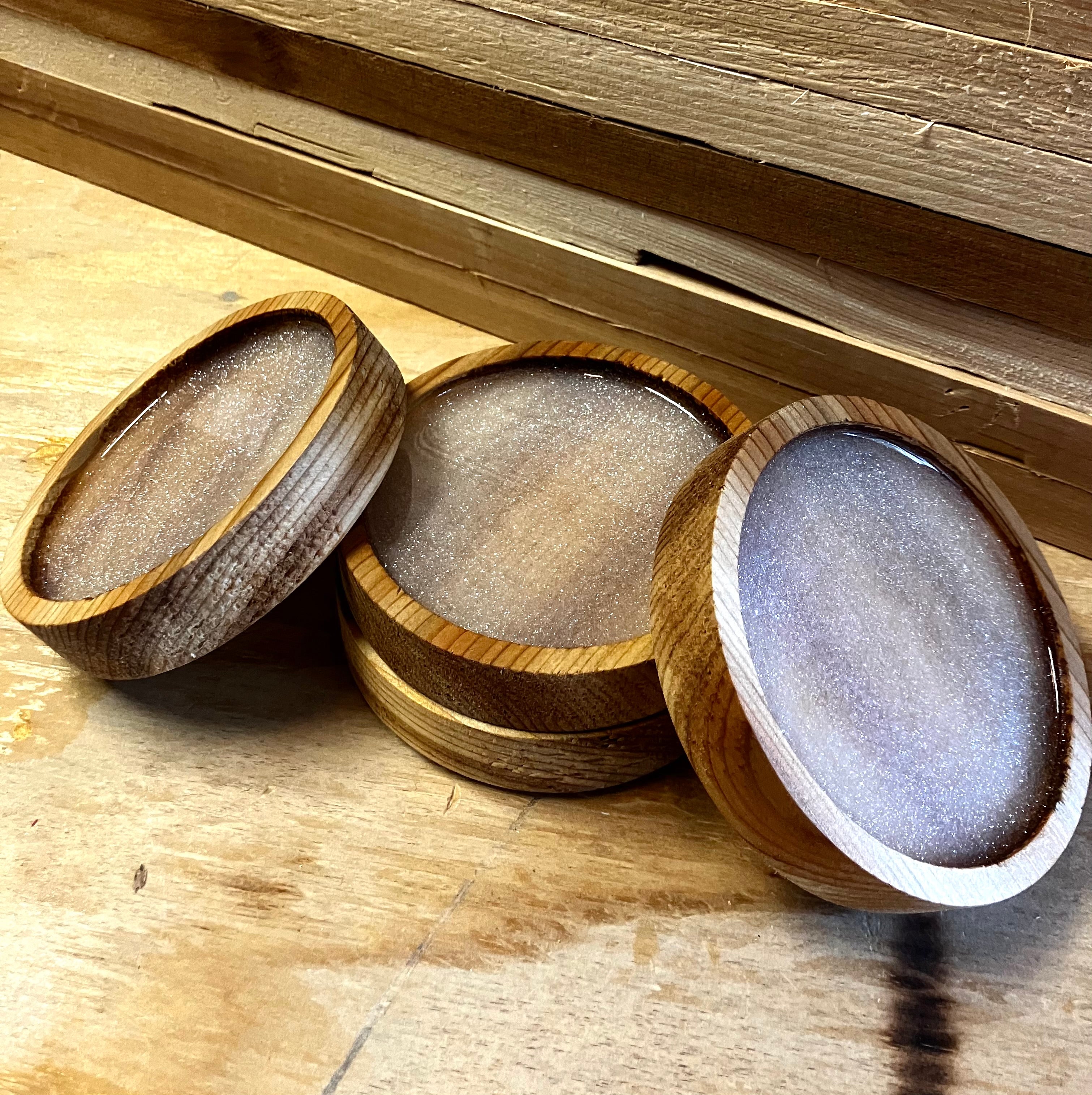 Displayed on a wooden table are three handcrafted wooden bowls with shimmering surfaces, accompanied by Willy's Woods' Customized Round Cedar Coasters. The bowls vary in wood tones and appear against stacks of wooden planks featuring customizable designs in the background.