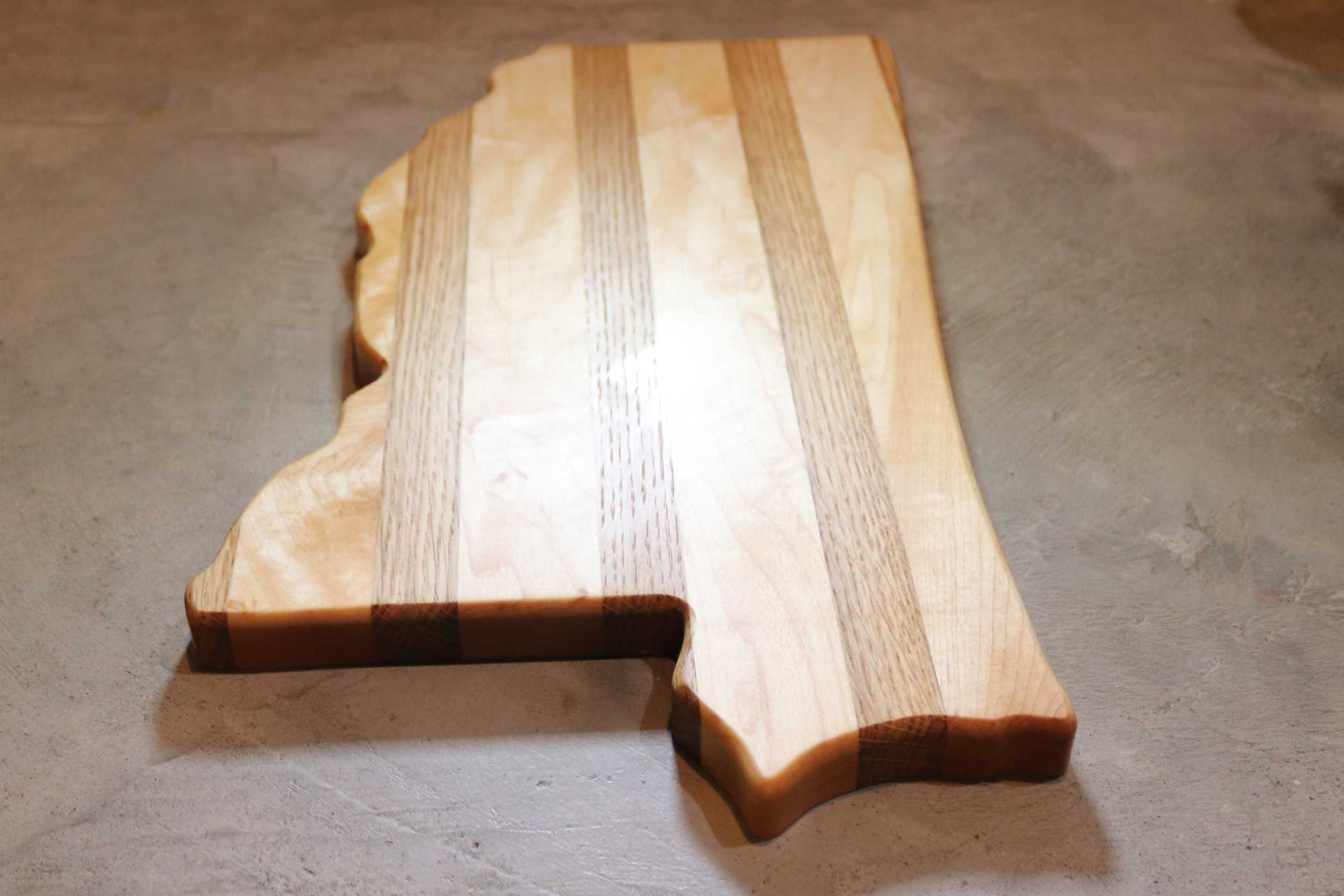 This My Store state-shaped cutting board artistically embodies Mississippi with alternating light and dark wood stripes, set against a textured gray surface.