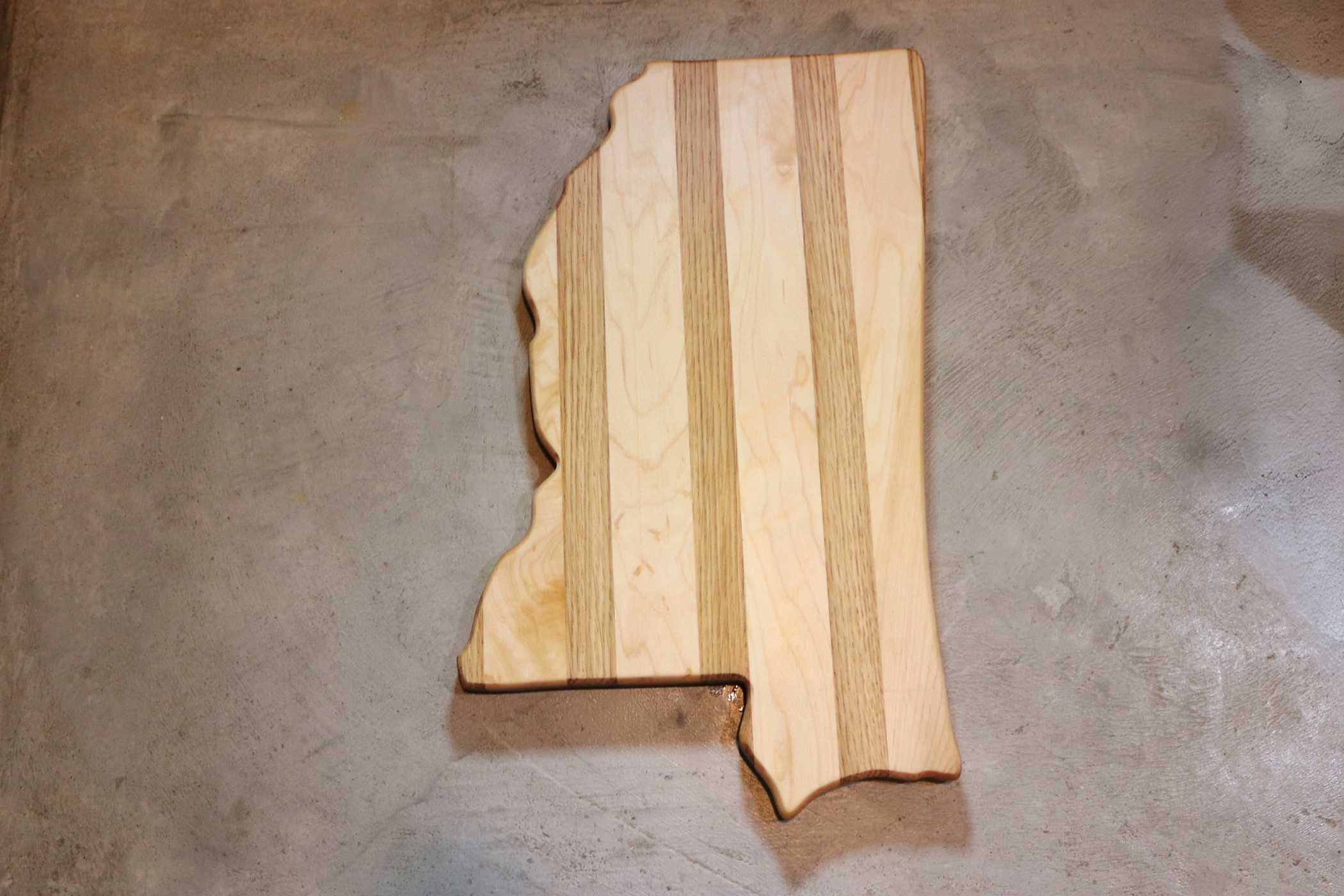 The State-Shaped Cutting Board by My Store, featuring premium craftsmanship with alternating light and dark wood strips in the shape of Mississippi, is set against a gray concrete background.