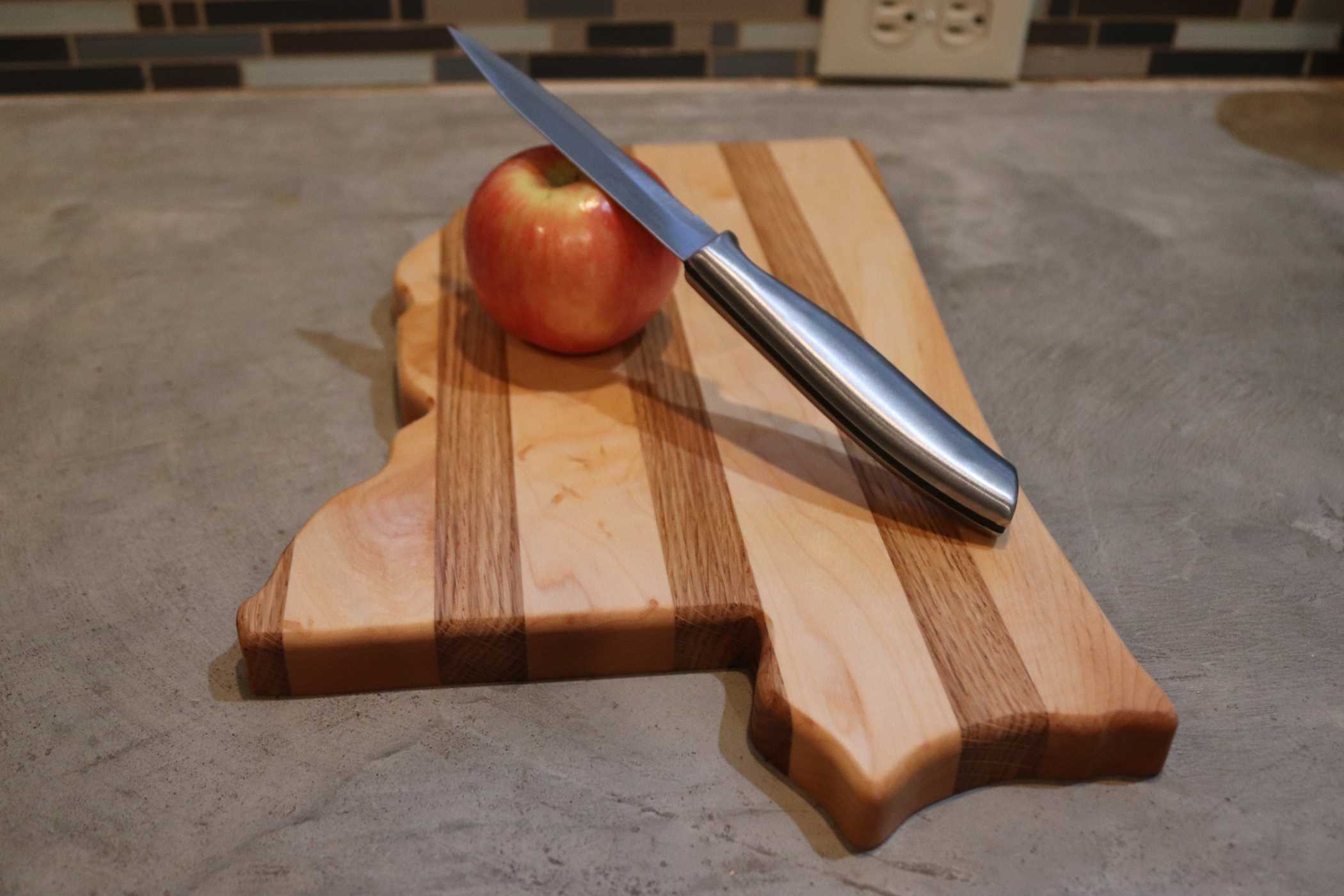 A Mississippi-shaped cutting board from My Store's State-Shaped Cutting Boards collection, featuring a striped wooden design, rests on the countertop. An apple and a sleek knife are placed on it, with an electrical outlet visible in the background.