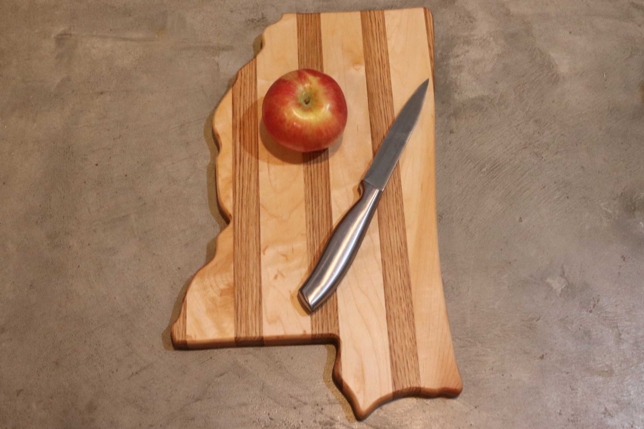 The State-Shaped Cutting Board by My Store, crafted with premium quality featuring lighter and darker wood stripes, holds a red apple and metal knife. This elegant custom piece rests on a gray surface, showcasing its unique design.