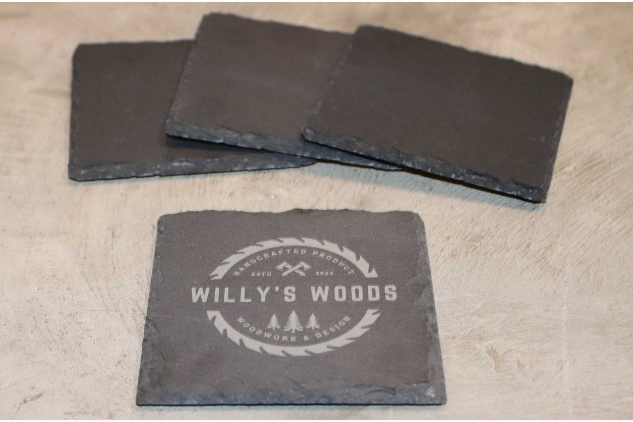 Four customizable slate square coasters from Willy's Woods are arranged on a light surface. The front coaster features the Willy's Woods logo with two trees and crossed tools, highlighting craftsmanship. A subtle clear coat enhances their durability and look.