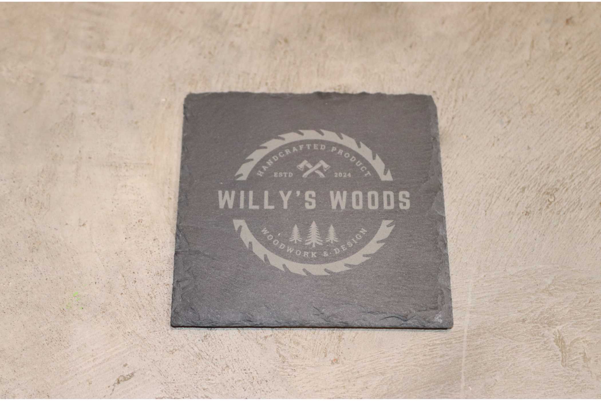 The Customizable Slate Square Coaster by Willy's Woods features an elegant engraving of "Willy's Woods" and a circular crossed saws logo. With "Handcrafted Products, Estd. 2022, Woodwork & Design," its design is enhanced by a clear coat against a textured, light background.