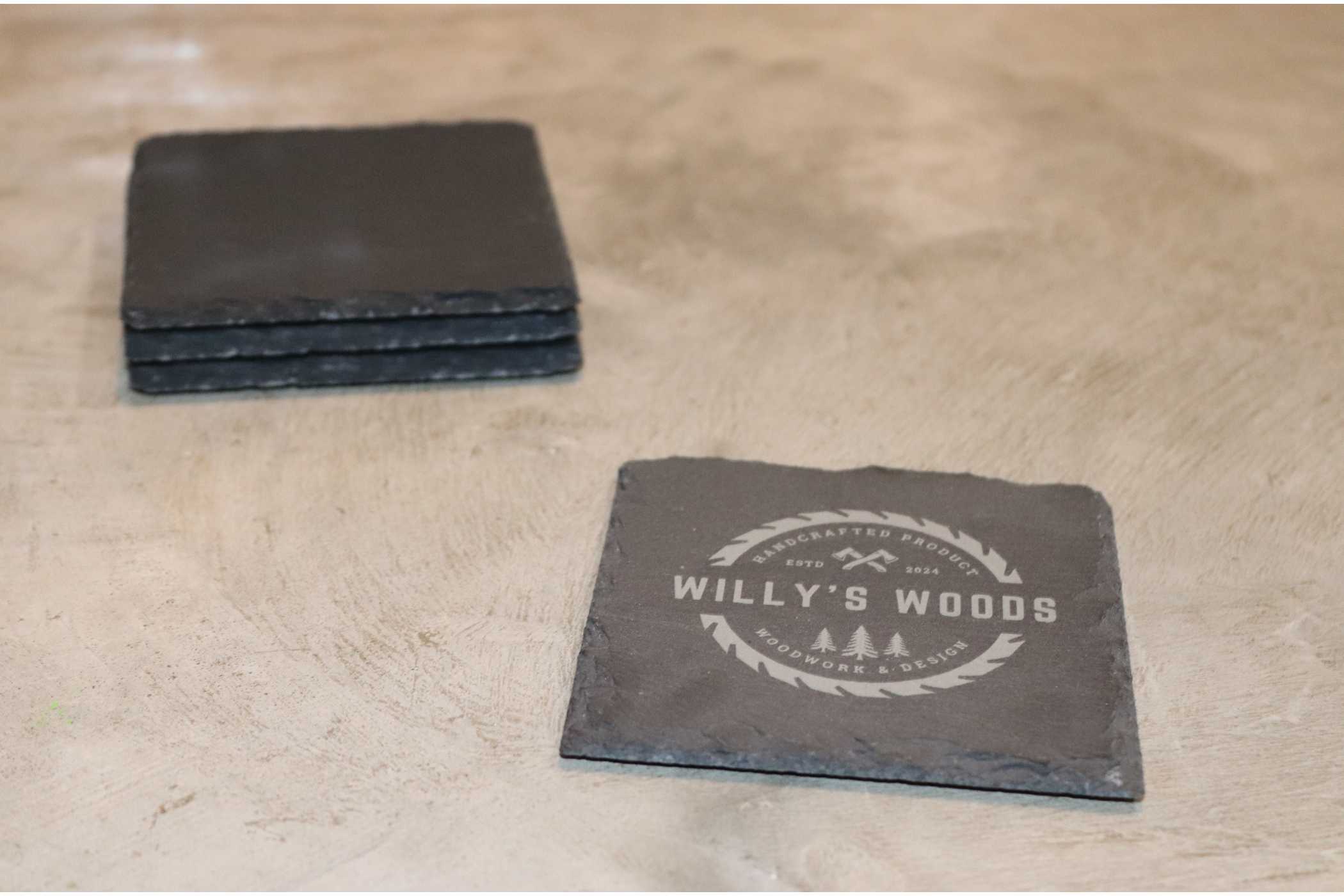 Four square black Customizable Slate Square Coasters from Willy's Woods are on a gray surface, three stacked to the left and one to the right showcasing a circular logo with "WILLY'S WOODS" and tree illustrations, all protected by a clear coat for durability.