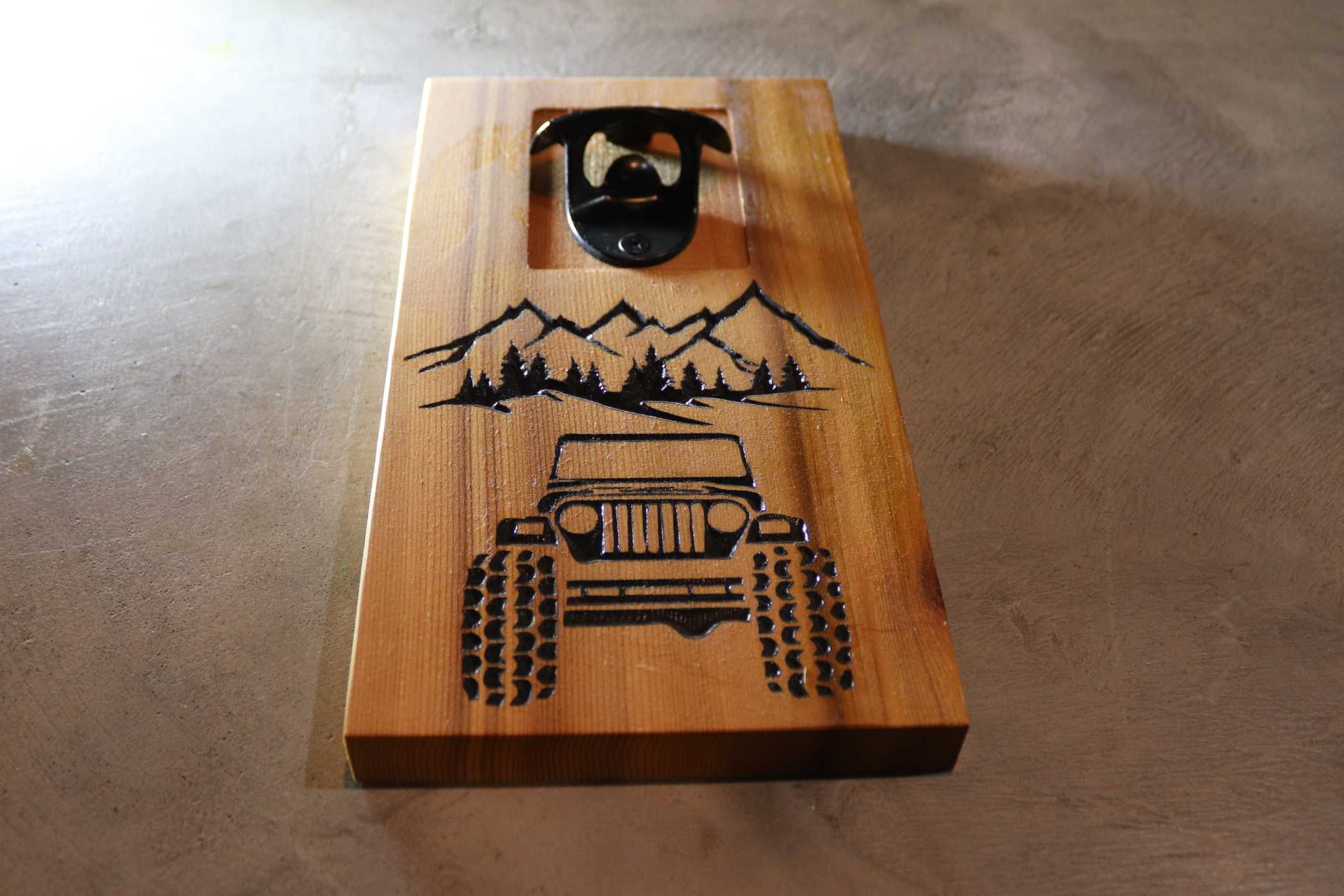 Willy's Woods presents the Customizable Cedar Wall Mounted Bottle Opener, featuring a metal top with custom engravings of a mountain scene and off-road vehicle, all on a gray base. Ideal for adding charm to any personalized home bar setup.