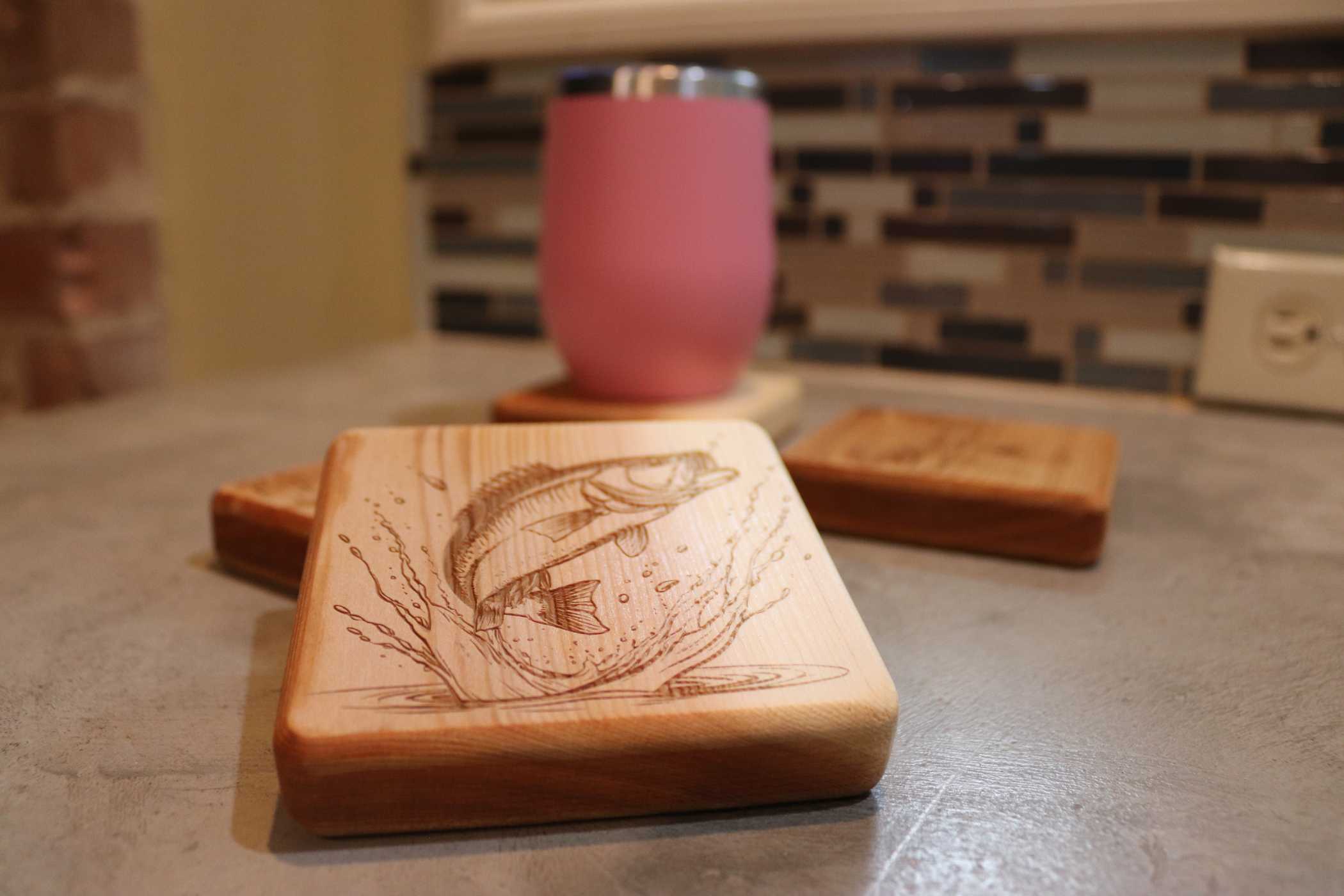 A Willy's Woods Customizable Cedar Square Coaster with an engraved fish design rests on the table, part of a charming set. More cedar coasters accompany a pink cup against a mosaic tile backsplash, with a power outlet visible in the background.