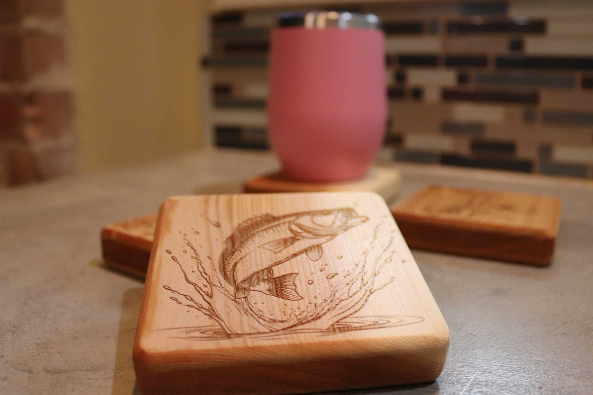 A table displays a customizable Cedar Square Coaster from Willy's Woods, featuring an engraved jumping fish design. In the background, additional coasters and a blurred pink mug with a silver rim are set against a neutral-colored tiled wall.