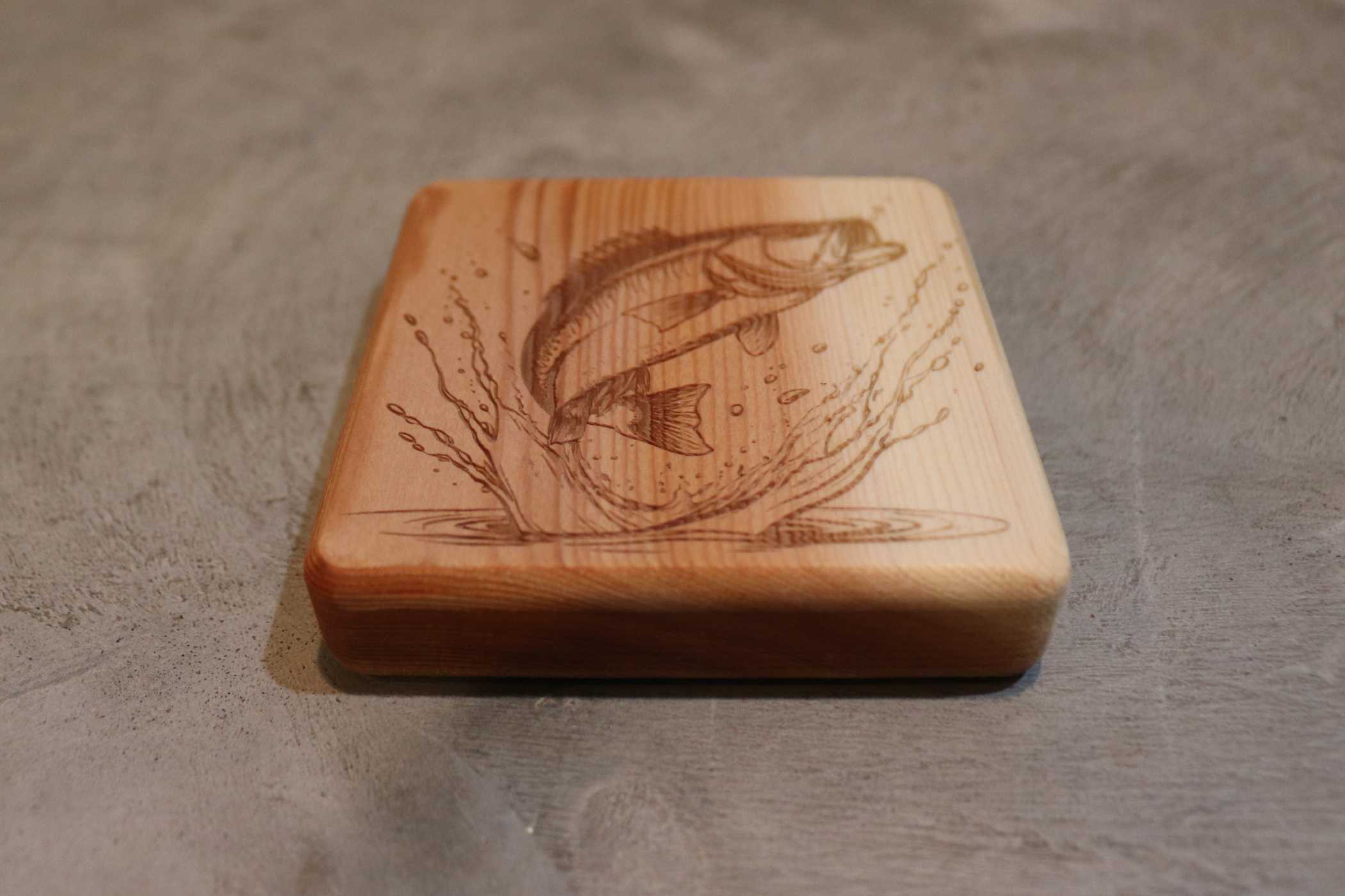 The Customizable Cedar Square Coaster by Willy's Woods features an engraved fish leaping with splashes, elegantly displayed on a textured gray surface, merging art and functionality.