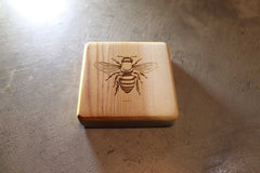 A square cedar coaster from Willy's Woods, engraved with a bee illustration, rests on a textured gray surface. Soft lighting emphasizes the intricate design. Part of the Customizable Cedar Square Coaster Set, it adds elegance to any home.