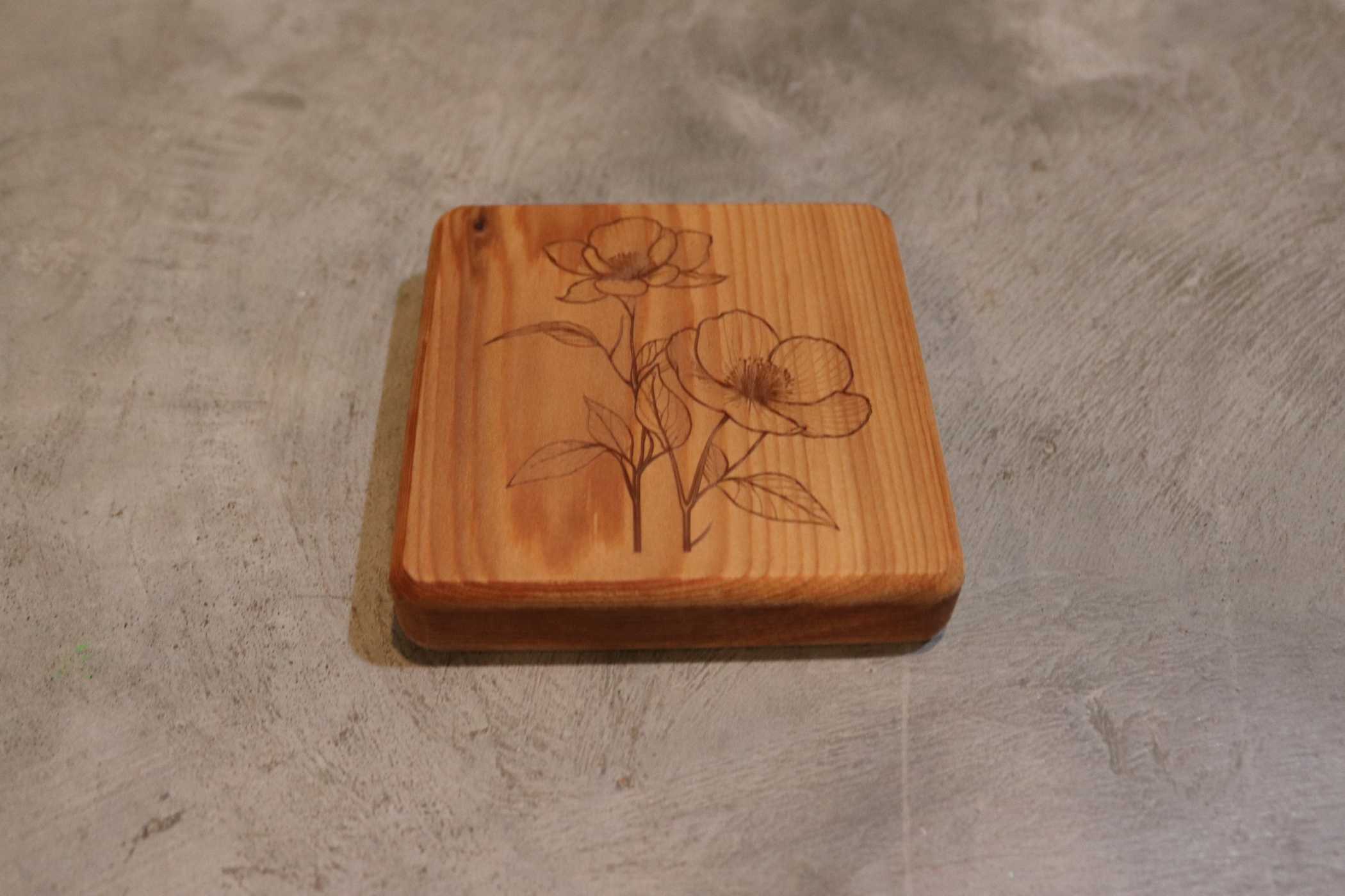 A coaster from Willy's Woods' Customizable Cedar Square Coaster Set features an engraved floral design with two blooming flowers and leaves, elegantly resting on a textured gray surface.