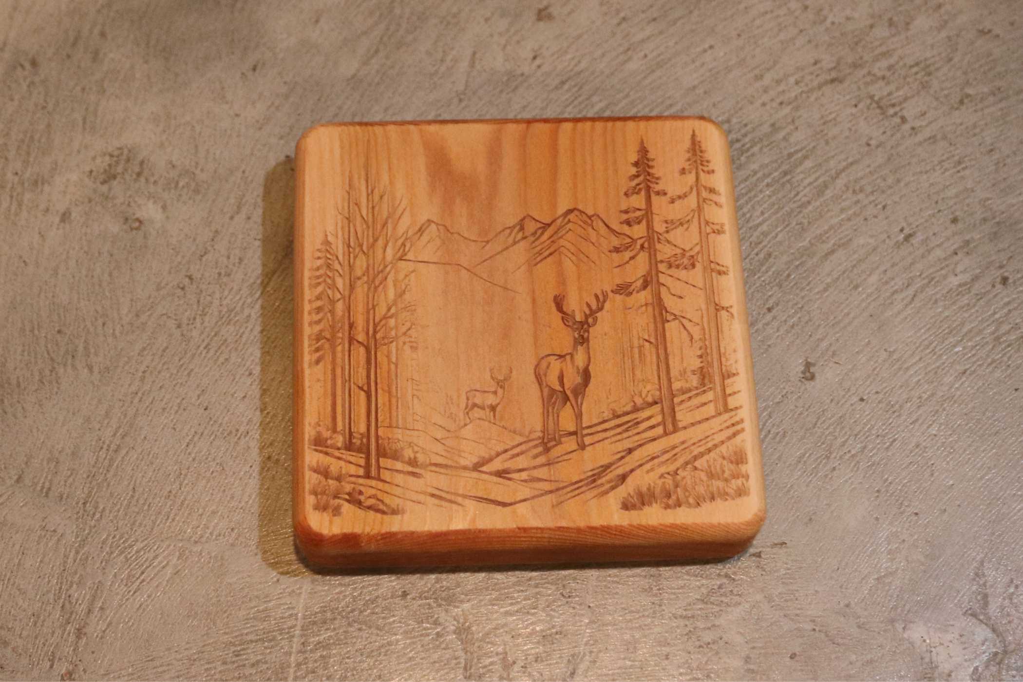 The Customizable Cedar Square Coaster Set by Willy's Woods includes square coasters engraved with a forest scene of two deer among trees and mountains, perfect for enhancing any rustic or natural-themed decor.