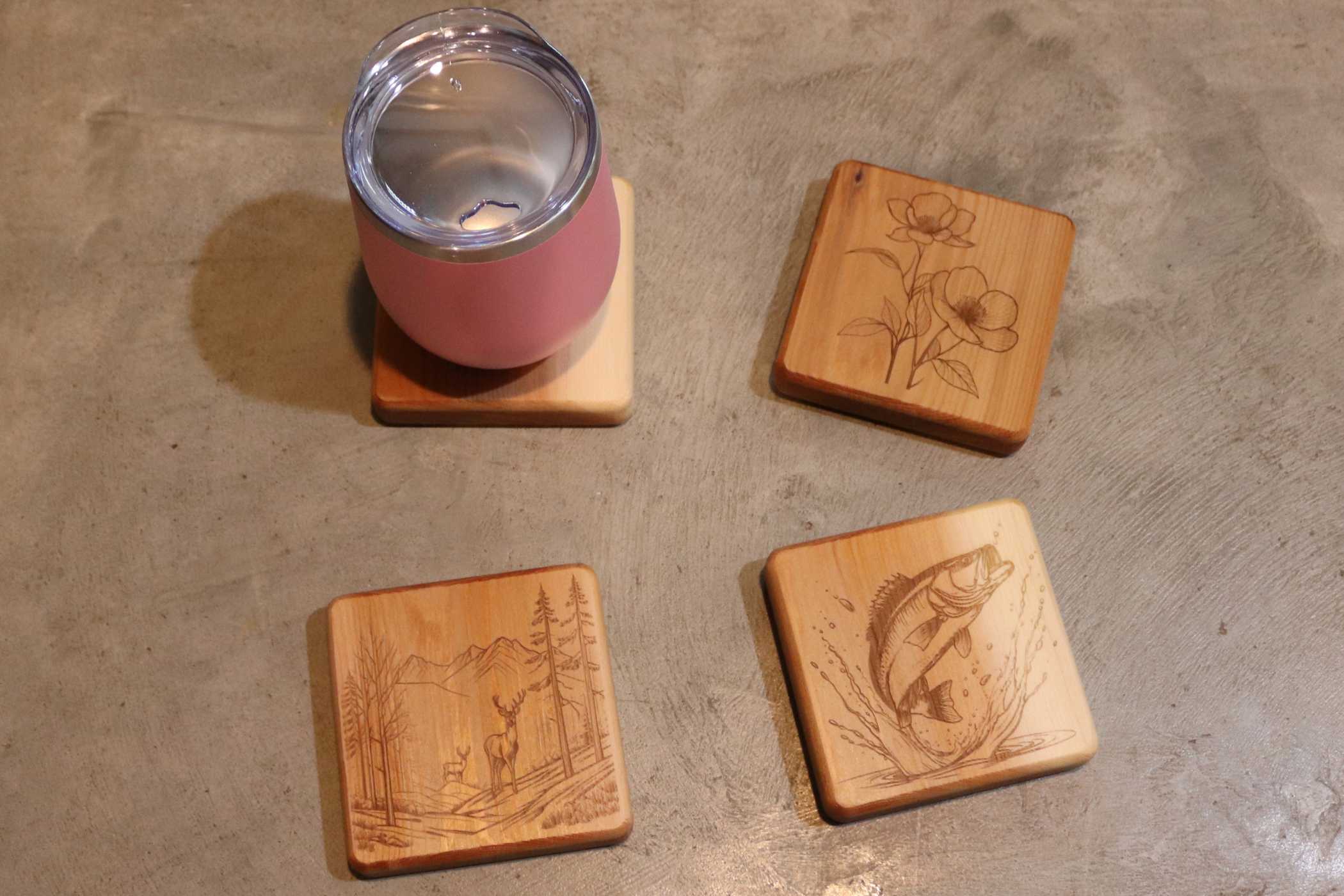 A pink tumbler with a lid rests on a wooden coaster. Nearby, Willy's Woods' Customizable Cedar Square Coaster Set features engraved designs: with flowers, mountains and deer, and a jumping fish. They are neatly arranged on a gray surface.