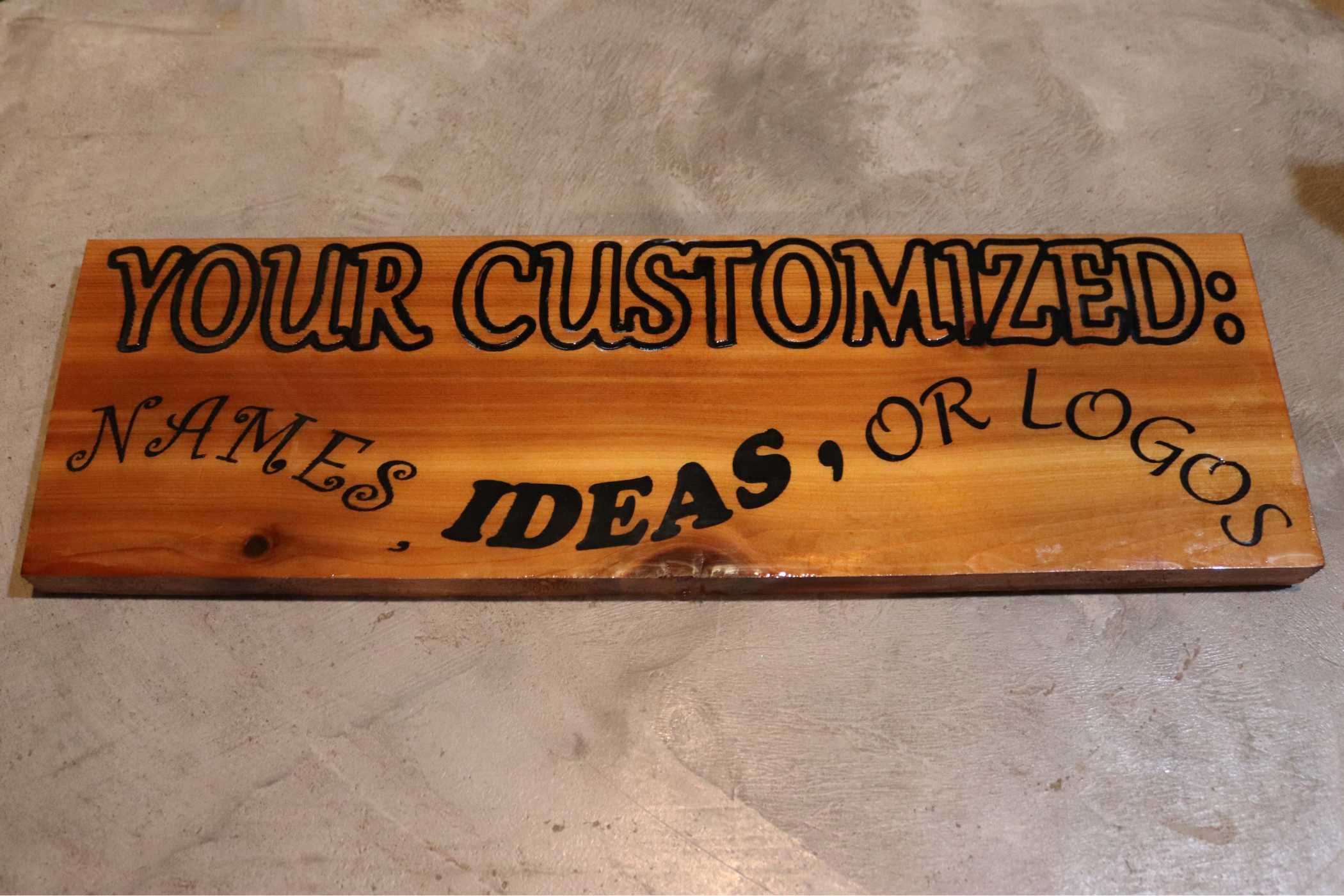 Willy's Woods offers Custom Cedar Wall Signs with bold black text for your customized names, ideas, or logos on light brown wood grain, elegantly displayed on a sleek gray concrete surface.