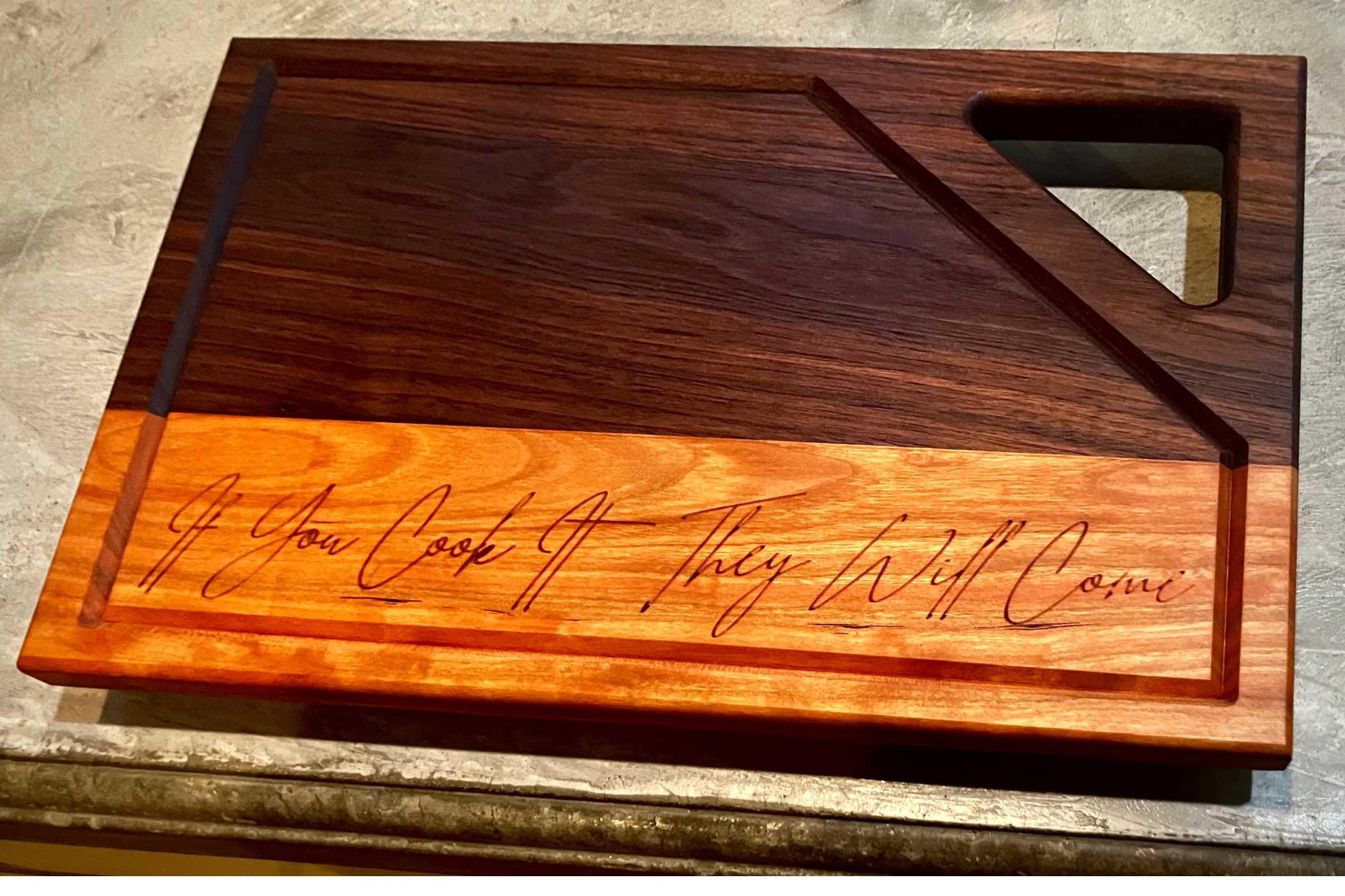 The Willy's Woods Customizable Walnut with Cherry Trim Wooden Cutting Board features a geometric design with a triangular handle, contrasting dark and light wood areas, and the engraving: "If You Cook It, They Will Come," making it ideal for any kitchen setting.