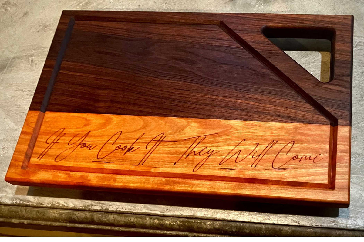 The Willy's Woods Customizable Walnut with Cherry Trim Wooden Cutting Board features a geometric design with a triangular handle, contrasting dark and light wood areas, and the engraving: "If You Cook It, They Will Come," making it ideal for any kitchen setting.