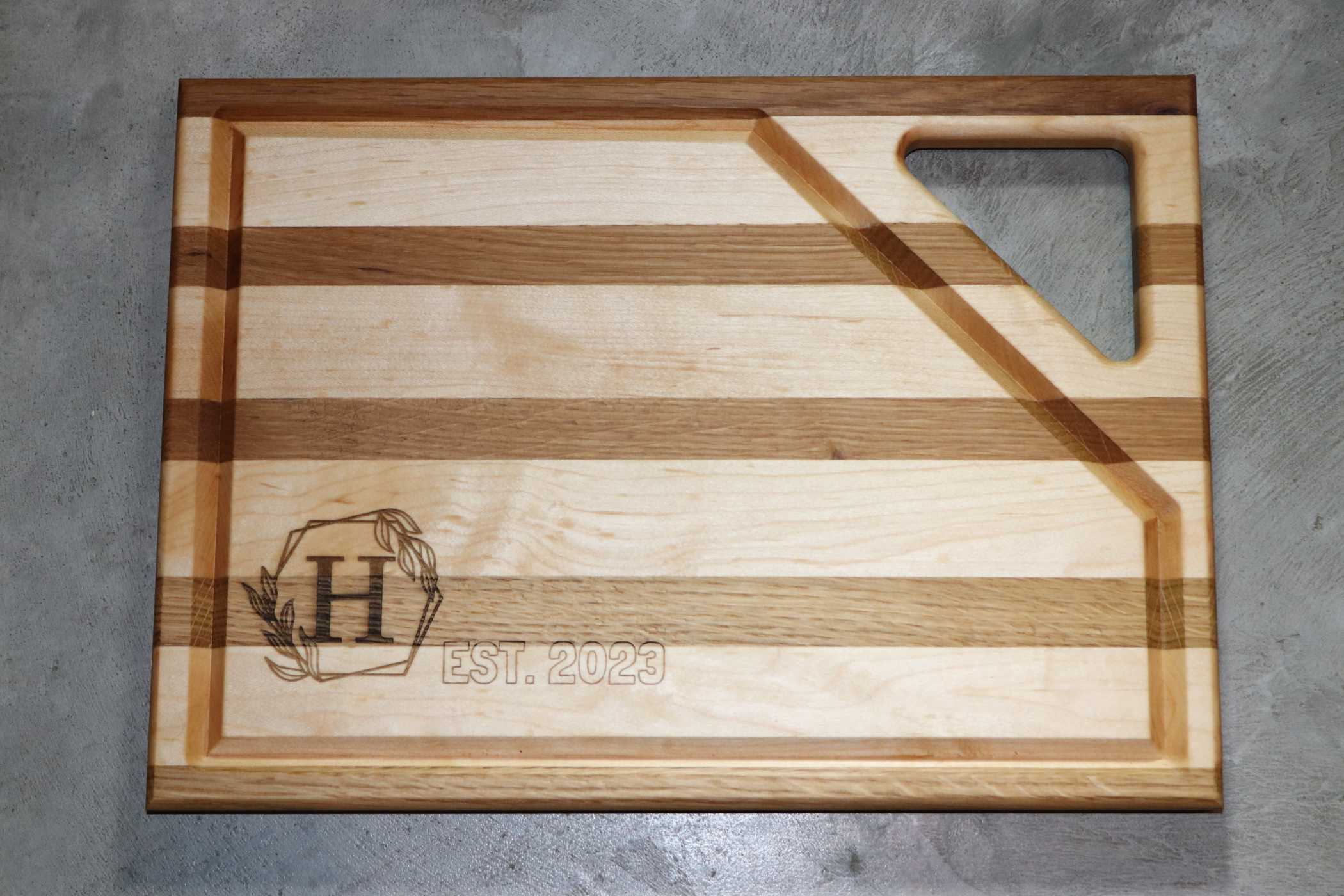 The Customizable Maple & White Oak Layered Wooden Cutting Board by Willy's Woods showcases alternating stripes and an engraved "H" with a wreath, plus "EST. 2023" text. A triangular cutout in the top right enhances its functionality and charm.