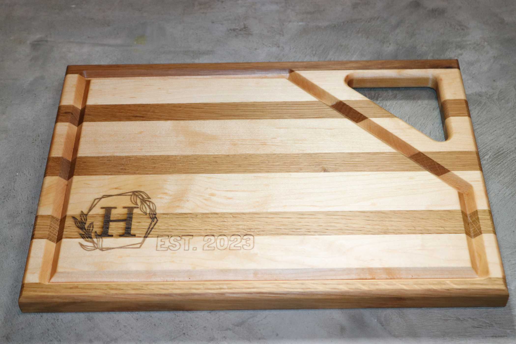 Willy's Woods presents a Customizable Walnut with Cherry Trim Wooden Cutting Board. It features an engraved letter "H" framed by a leaf motif and "EST. 2023," plus a top-right carved handle, offering elegance and custom craftsmanship to any kitchen.