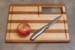 On a gray surface lies Willy's Woods' Customizable Walnut with Cherry Trim Wooden Cutting Board, featuring horizontal stripes. A red apple sits to the left, and a stainless steel knife is on the right. The board's rectangular cutout adds charm, making it perfect for any kitchen setting.