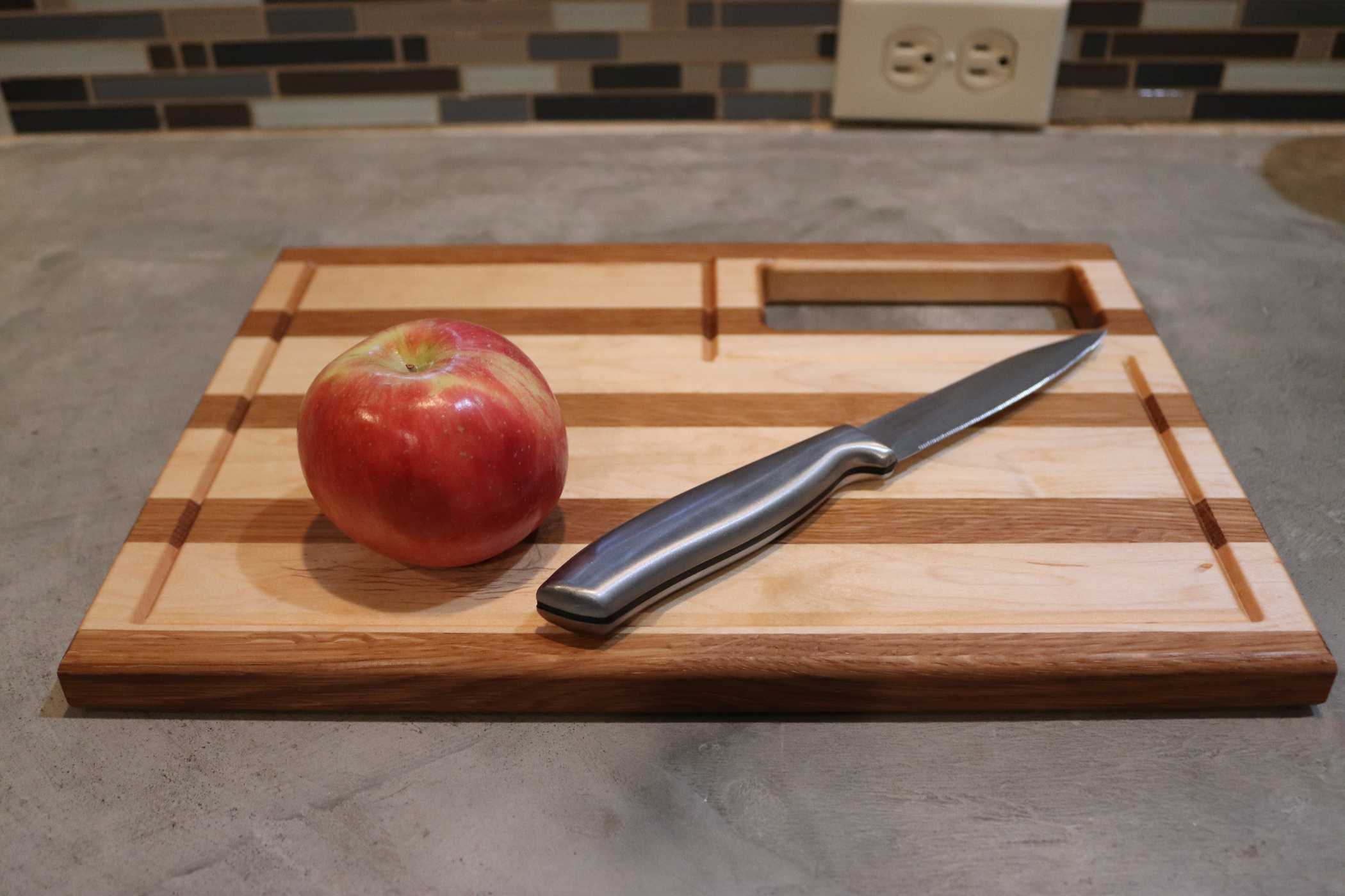 A red apple and a silver knife rest on Willy's Woods' Customizable Walnut with Cherry Trim Wooden Cutting Board. This handcrafted board features unique designs and lies on a gray countertop, with a tiled backsplash and dual electrical outlet in the background.