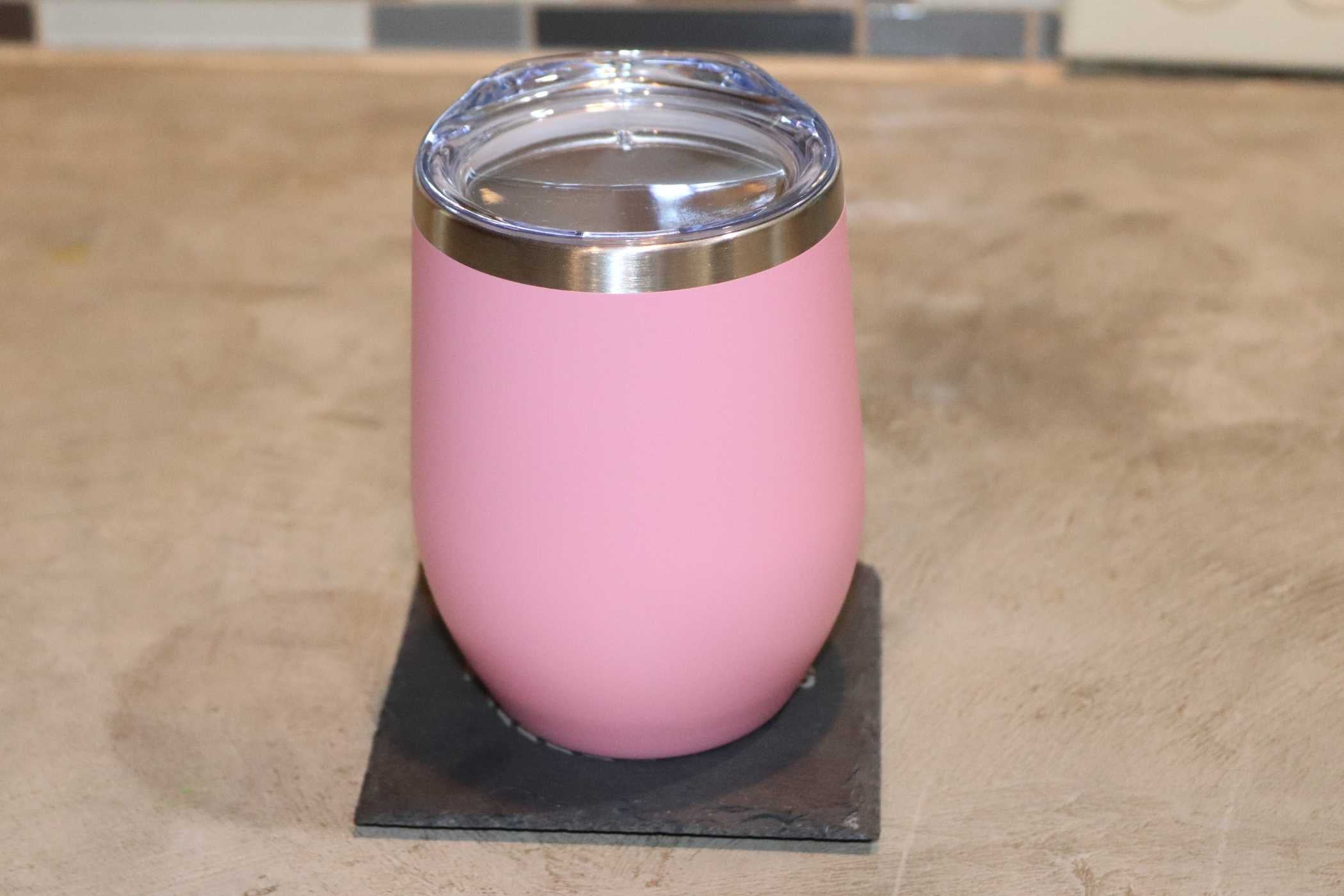 A Willy's Woods custom engraved pink tumbler with a metallic rim and clear lid sits on a dark coaster on a beige surface, set against an elegant tiled wall.